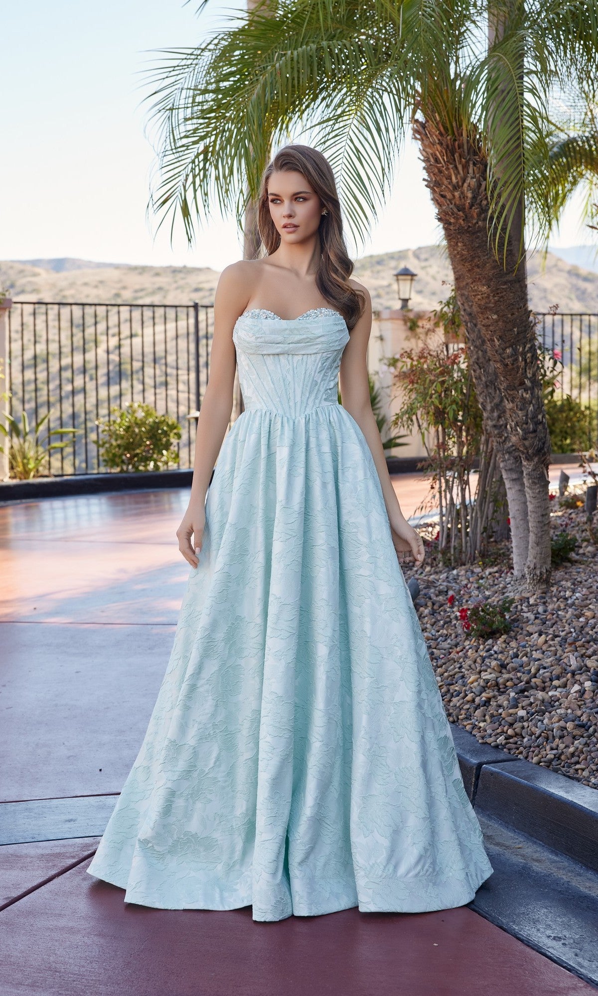 Outdoors by palm trees, a woman looks to the side while wearing the Nox Anabel T1912 long prom dress, featuring a strapless design and a textured skirt.